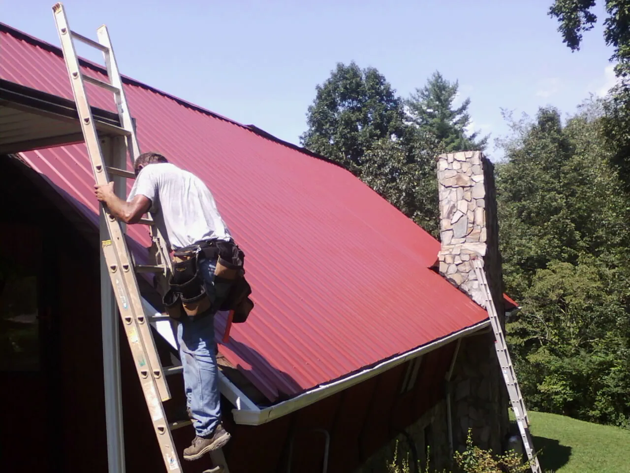 Roofer on a ladder beside a red metal roof and stone chimney — Soffit & Fascia Repair by Roofing Brothers in Killduff